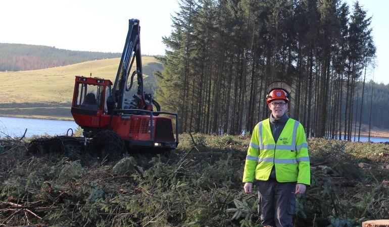 Man in a high visibility jacket stands in front of a digger and a small group of trees Man in a high visibility jacket stands in front of a digger and a small group of trees