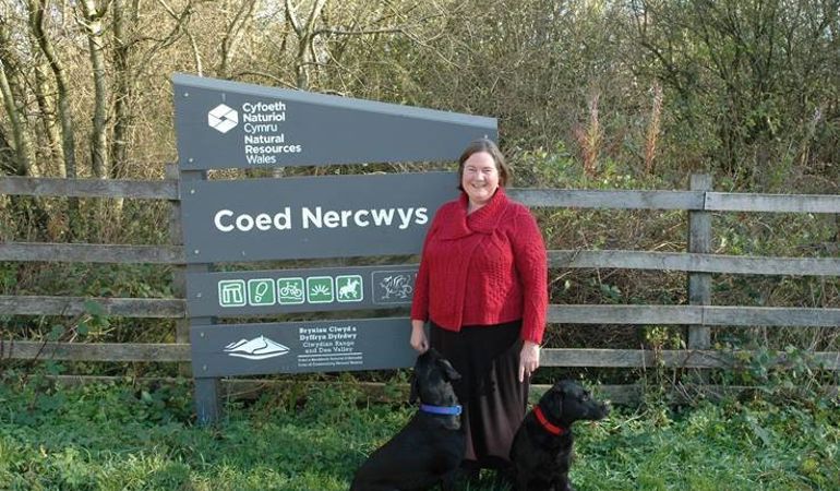 Clare Pillman CEO, wearing red jacket stood in front of the NRW Coed Nercwys sign with two black labrador dogs Clare Pillman CEO, wearing red jacket stood in front of the NRW Coed Nercwys sign with two black labrador dogs