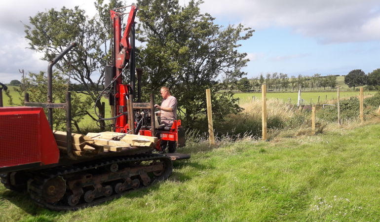 Ffotograff o ddyn yn gyrru peiriant ffensio fawr wrth ymyl Afon Wygyr gyda pyst newydd eu gsod y tu ôl iddo Ffotograff o ddyn yn gyrru peiriant ffensio fawr wrth ymyl Afon Wygyr gyda pyst newydd eu gsod y tu ôl iddo